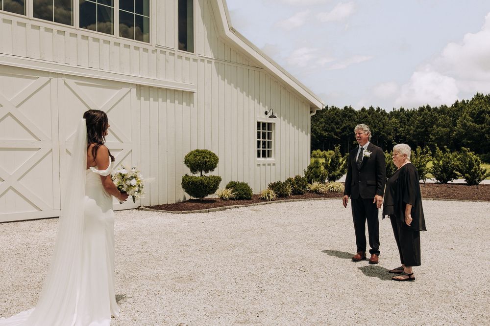 Bride in fitted white gown shares first look moment with parents outside white barn venue.