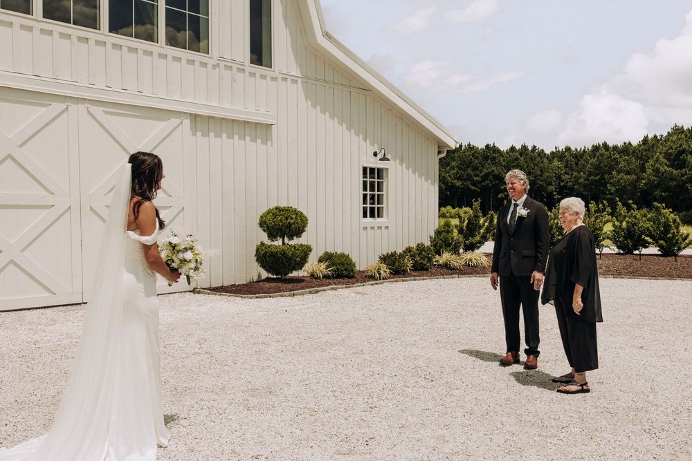 Bride holding white floral bouquet during outdoor first look with parents at barn venue.