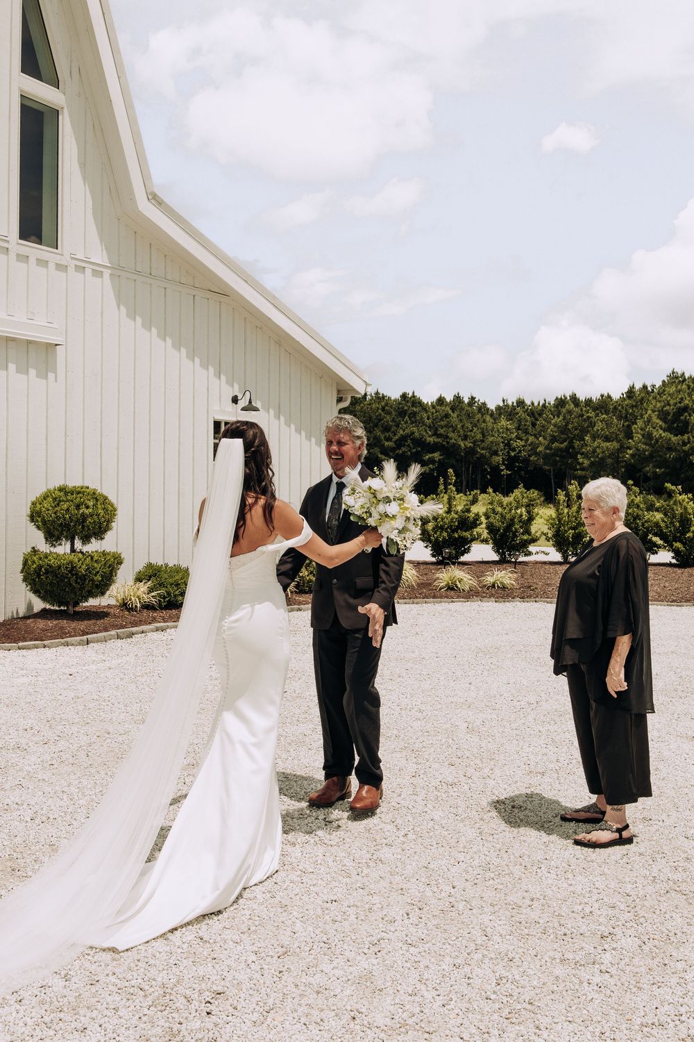Bride in white gown with veil meets older couple outside white barn venue on sunny wedding day.