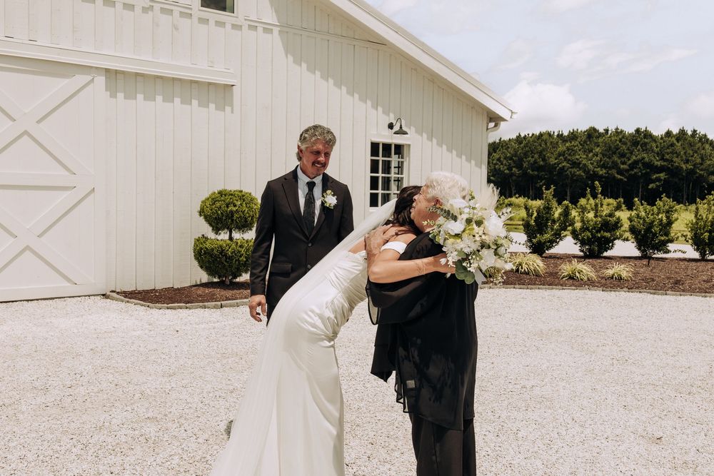 Bride in fitted white gown hugs guest while groom smiles in background outside white barn venue.