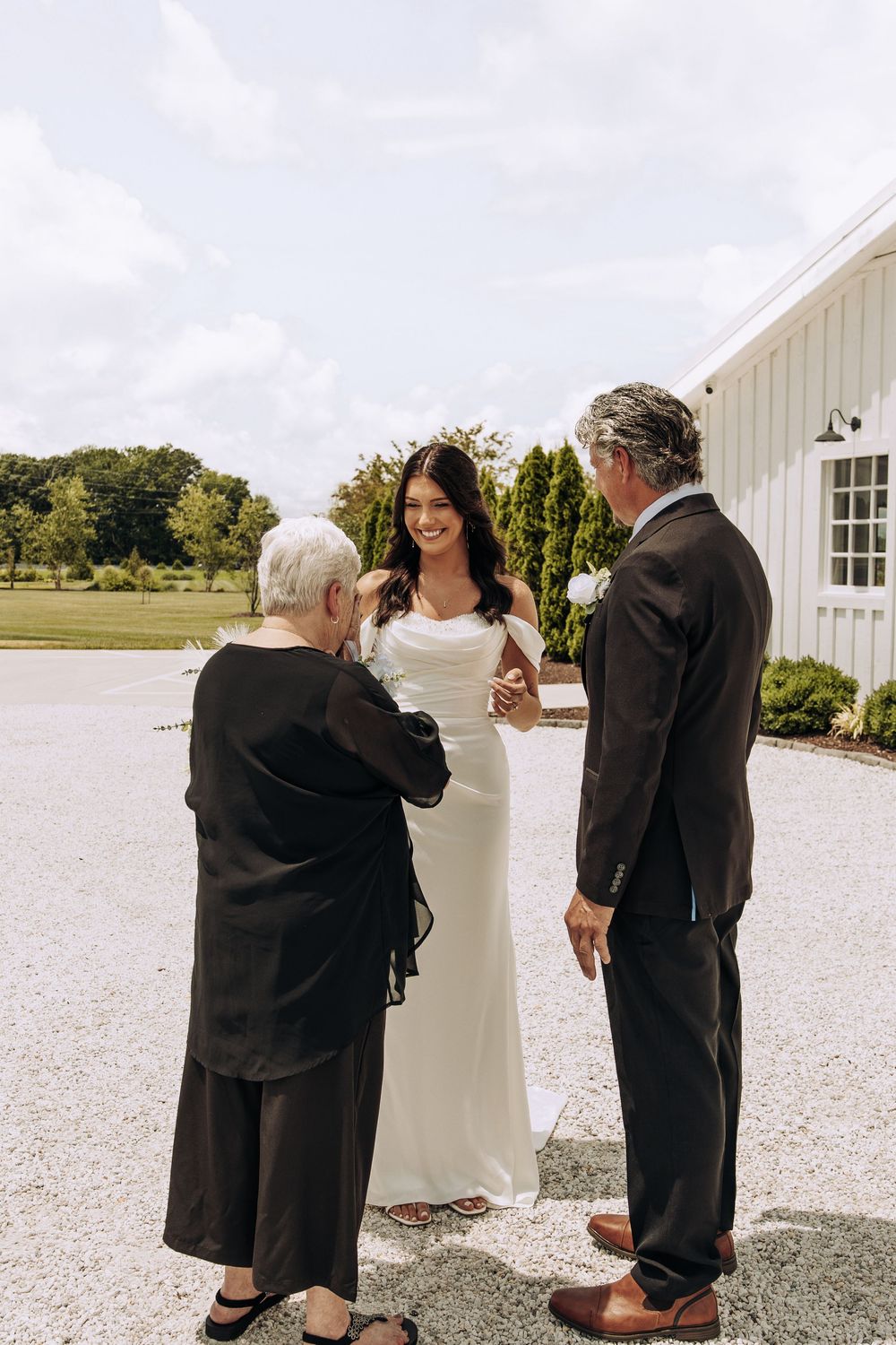 Smiling bride in white satin gown holds hands with older couple outside white barn wedding venue.