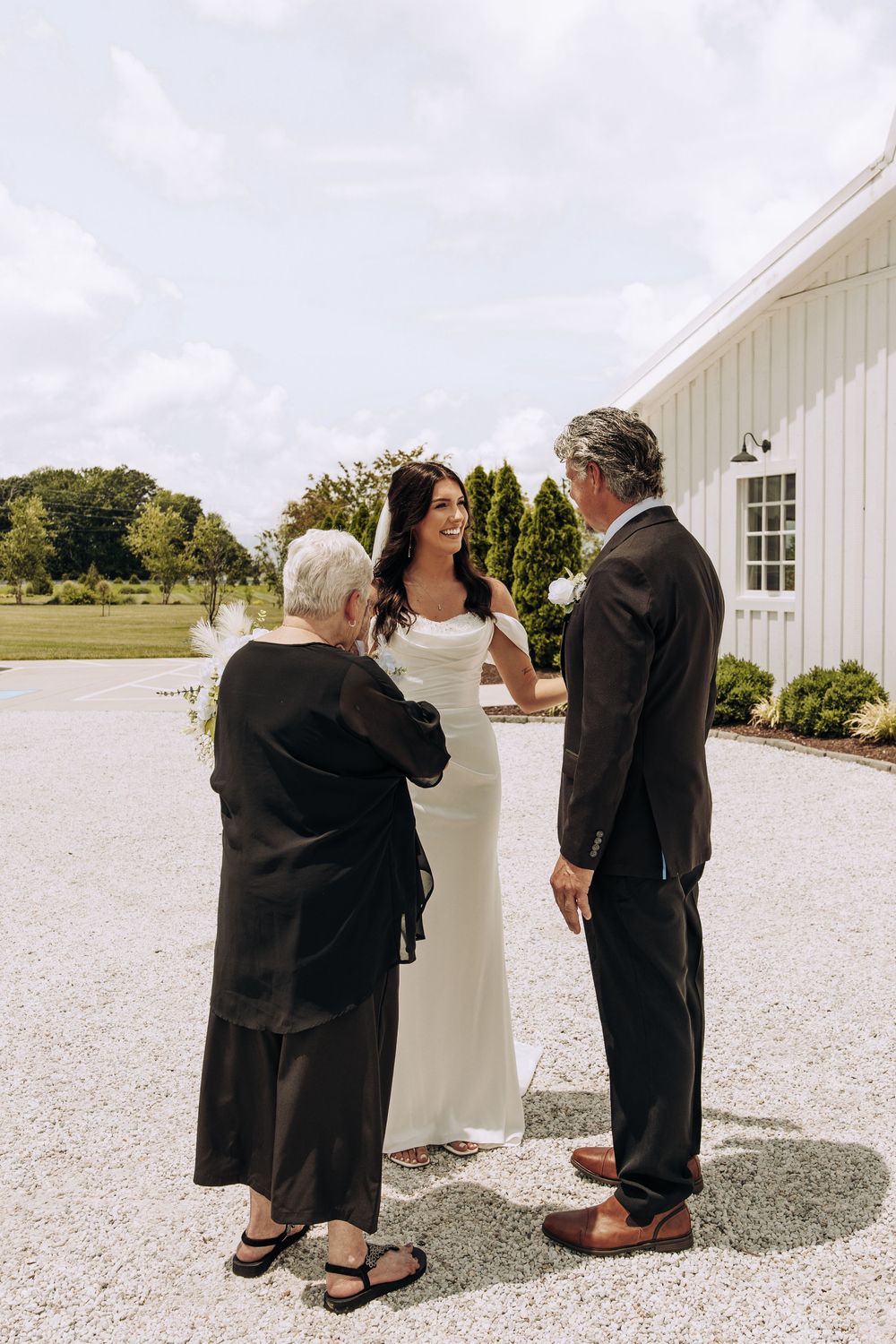 Bride in elegant white gown laughs with elderly woman and man in suit outside white barn venue.