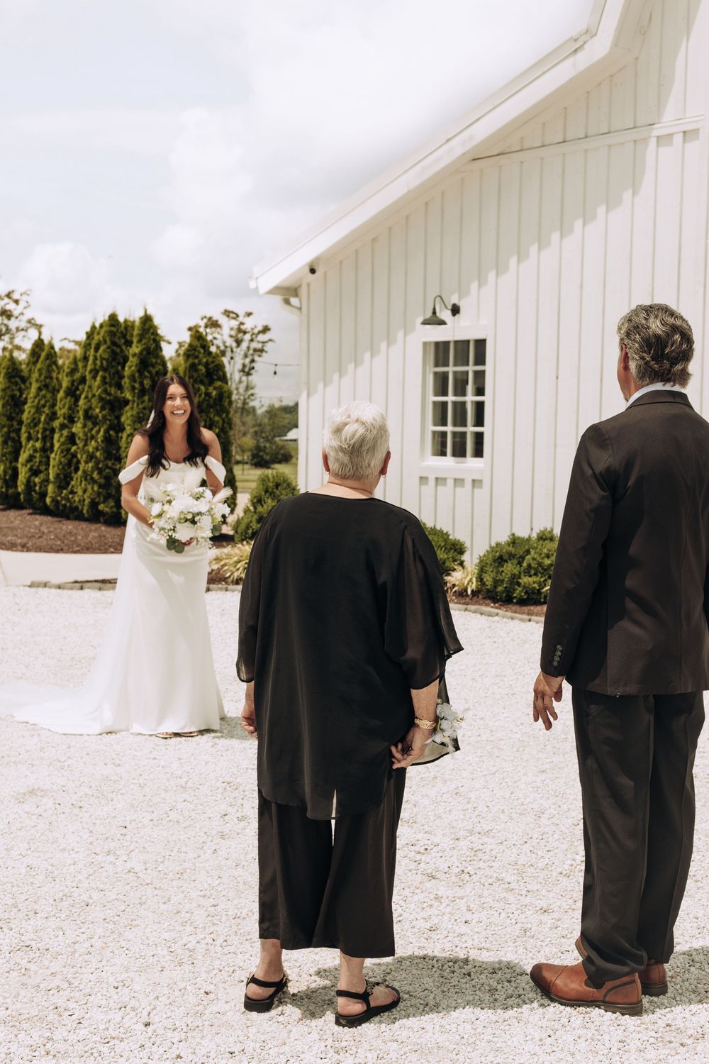 Smiling bride holding white bouquet greets parents during first look at white barn venue.
