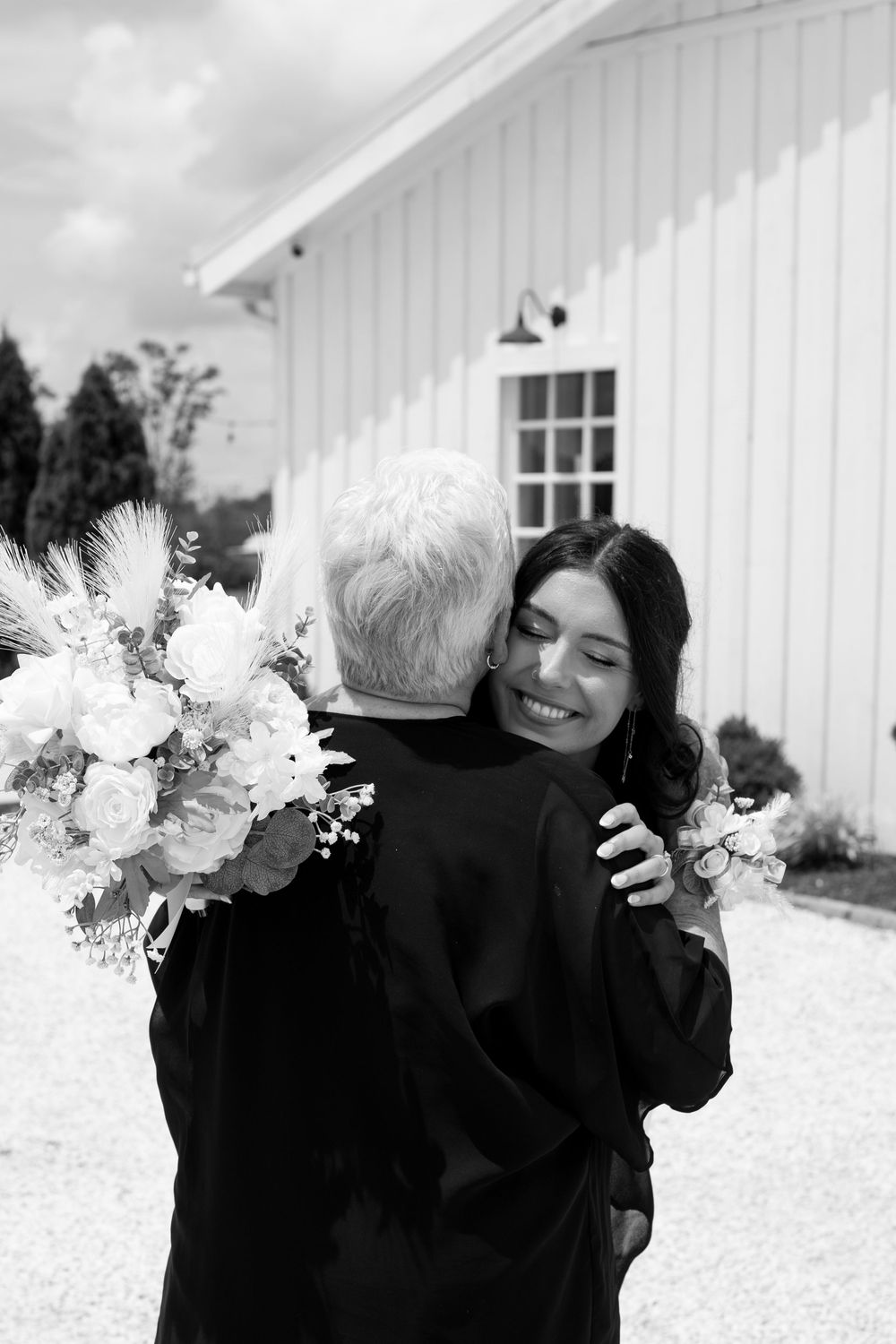 Black and white photo of smiling dark-haired bride hugging elderly woman holding white wedding bouquet.