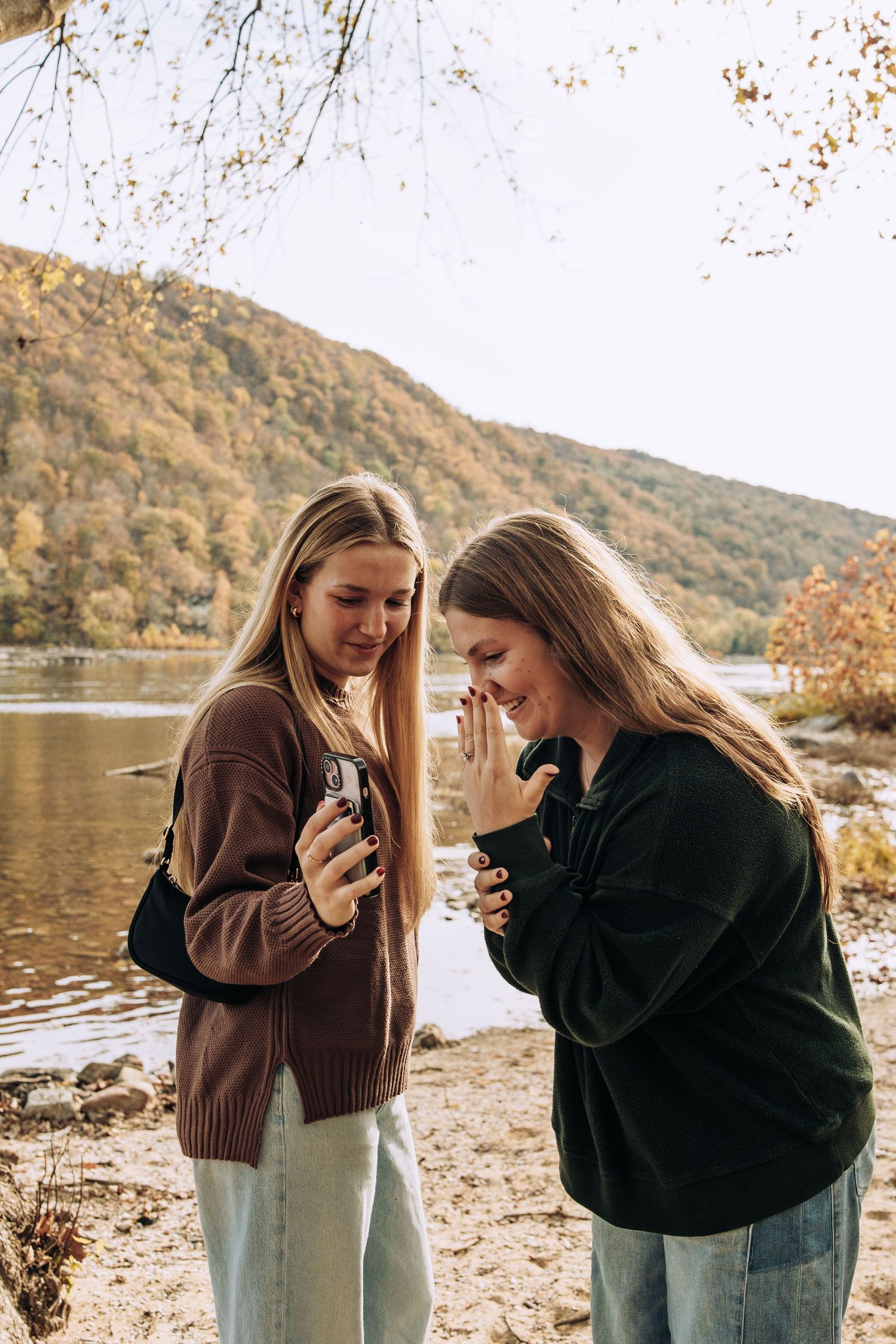Two young women laugh together while looking at a camera by a scenic autumn river.
