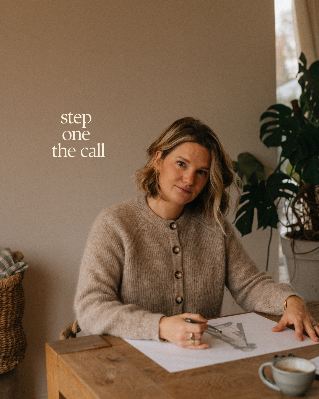 A person in a beige sweater sits at a wooden desk with a coffee cup and papers, working in a cozy home setting.