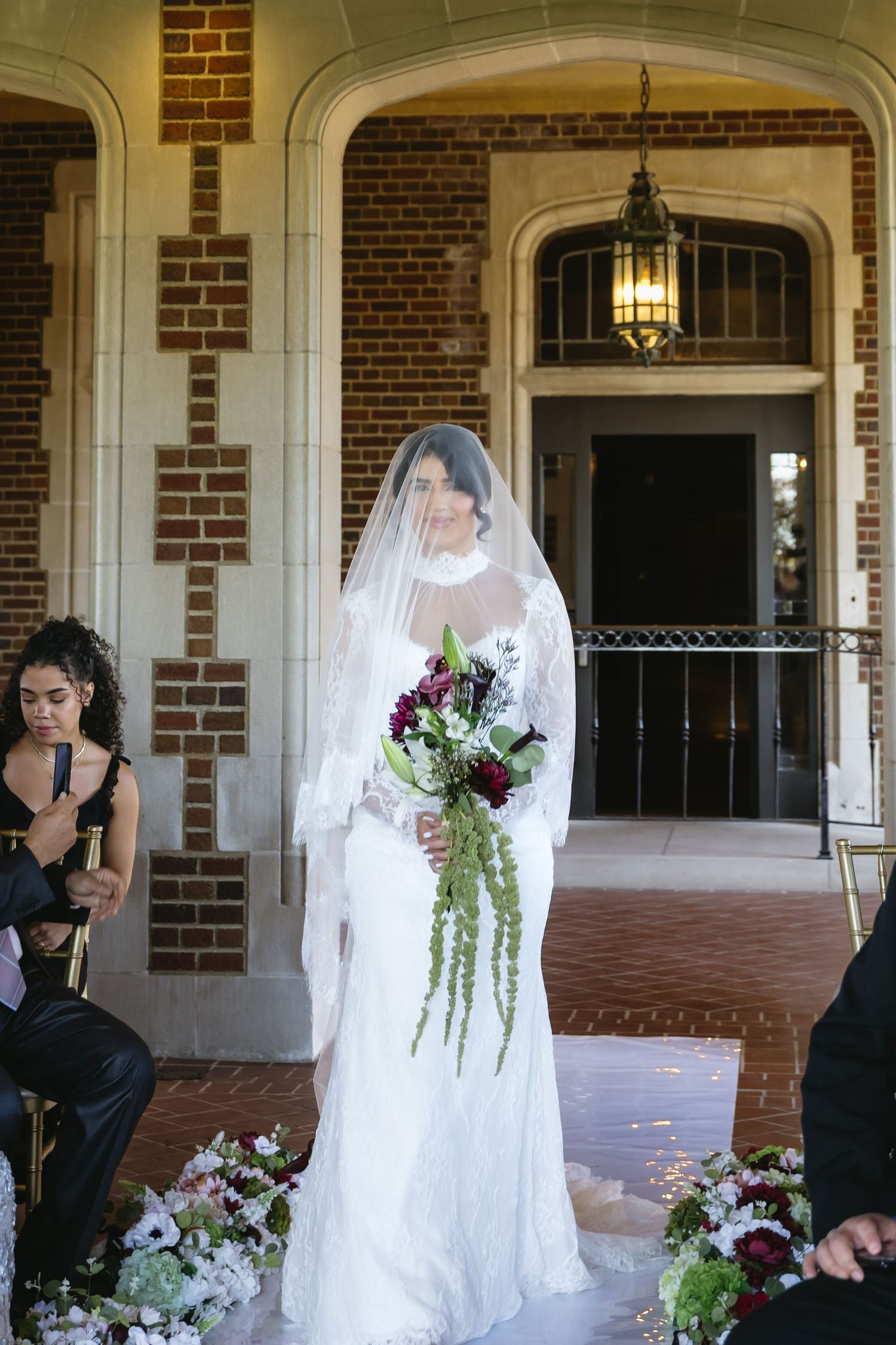 A bride in a white wedding dress poses for photos on a brick porch with floral arrangements and columns.