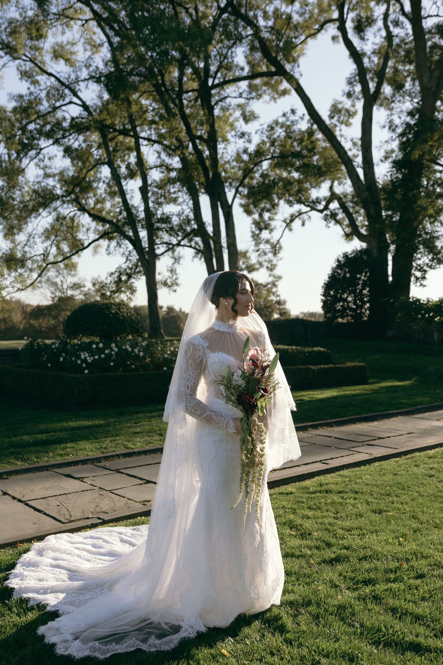Two people in white wedding attire stand together on a grassy lawn with tall trees in the background.