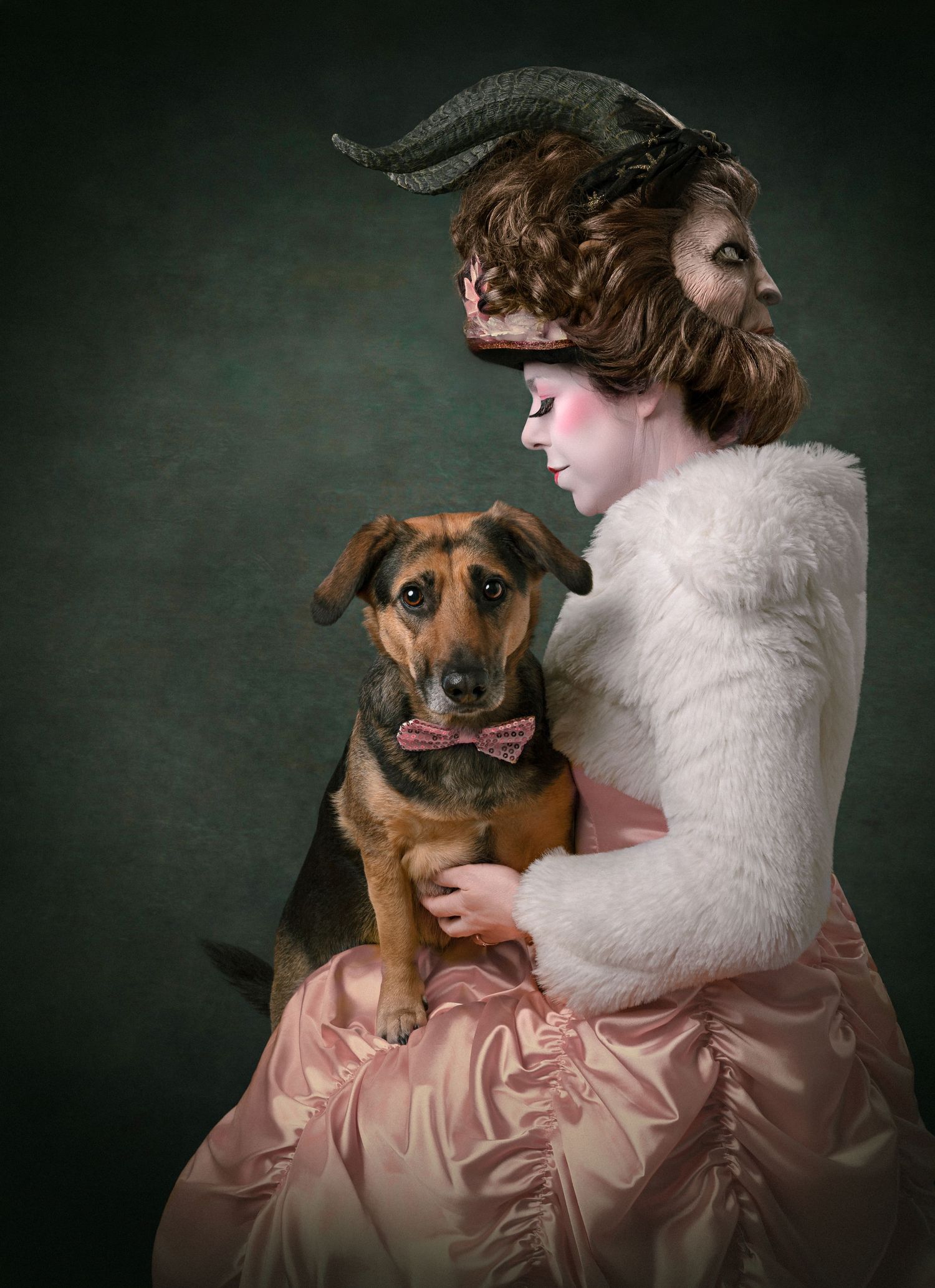 A brown dog poses with someone wearing a pink silk dress and white fur shawl against a dark background. fine art hondenfoto in de studio