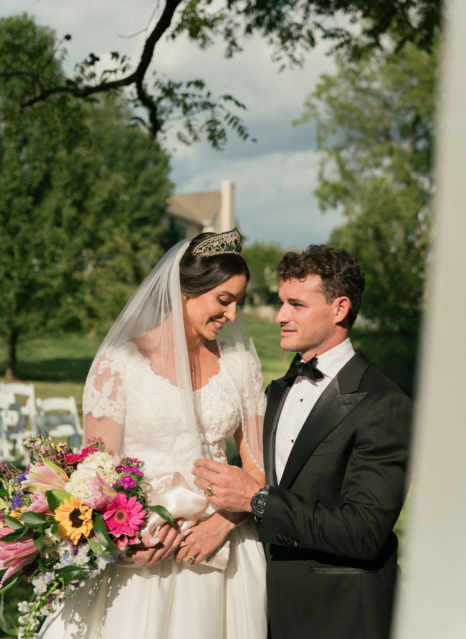 A wedding couple shares a tender moment near white columns in an outdoor garden setting.