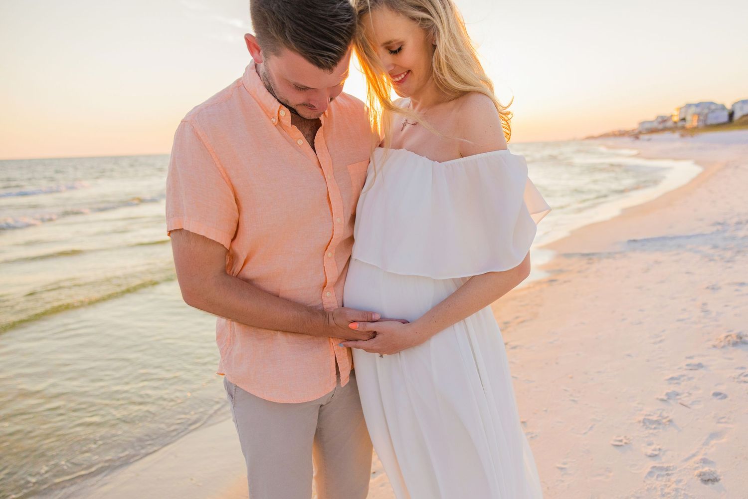 A couple shares an intimate moment on a sandy beach at sunset wearing soft pink and white outfits.