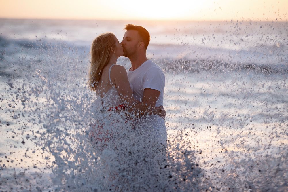 Couple kissing as waves crash behind them at sunset - Emotional couple portraits by photoFuerte