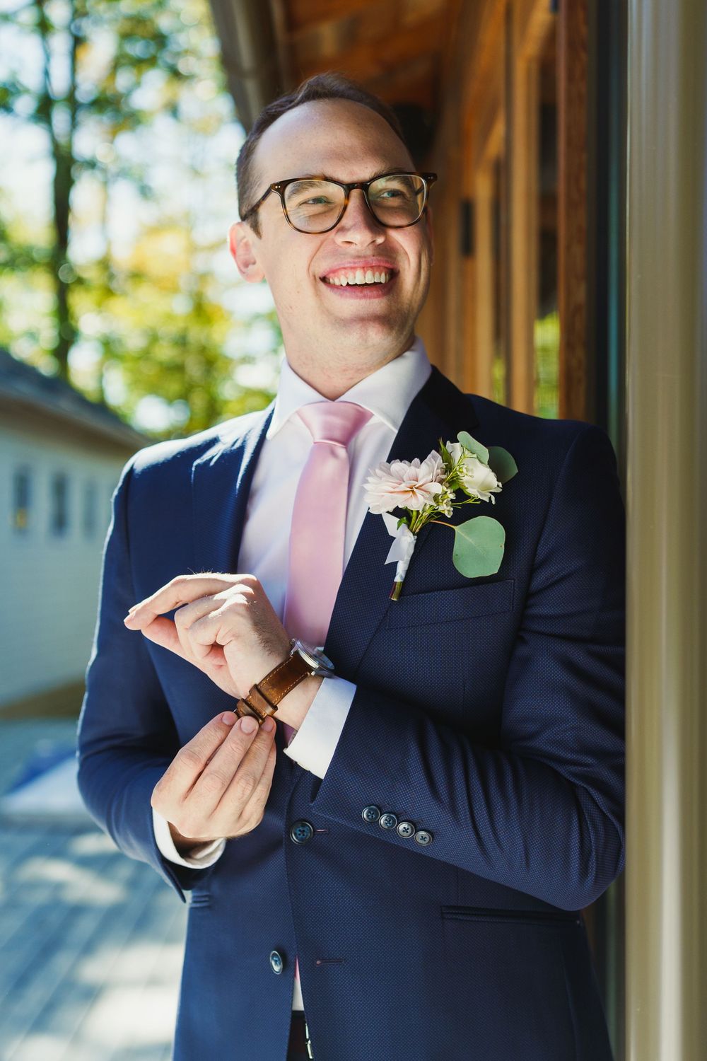 A groom in a navy suit and pink tie adjusts his cufflinks while smiling.