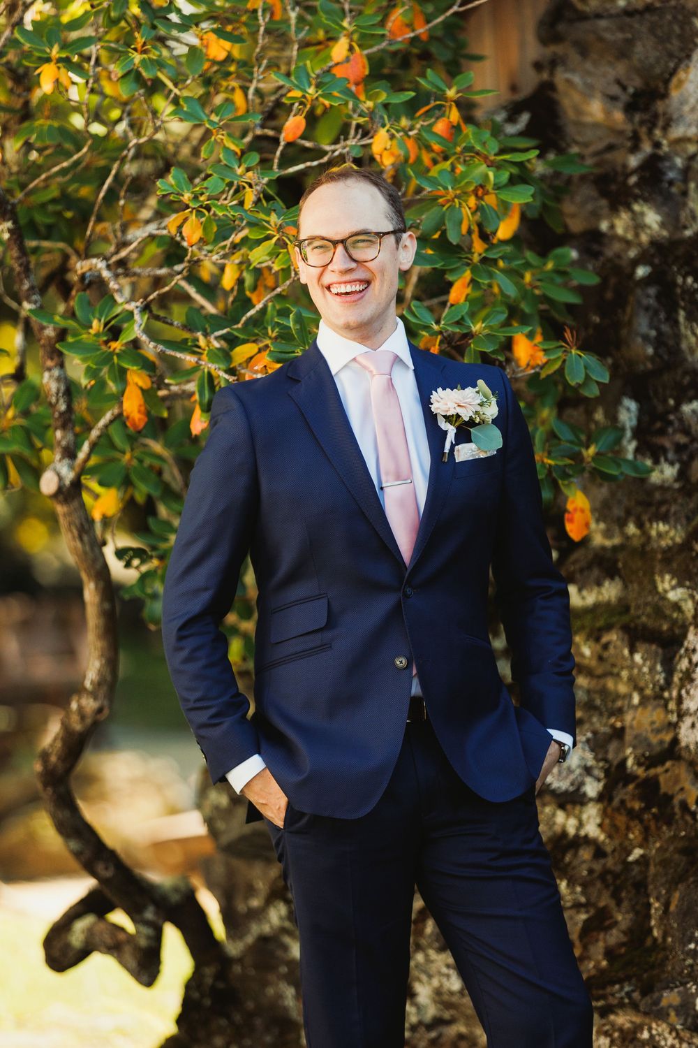 A groom in a navy suit and pink tie poses in front of orange blossoms.