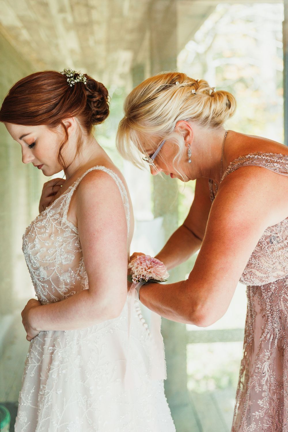 A tender moment as a wedding dress is being fastened in a sunlit room with delicate lace details.