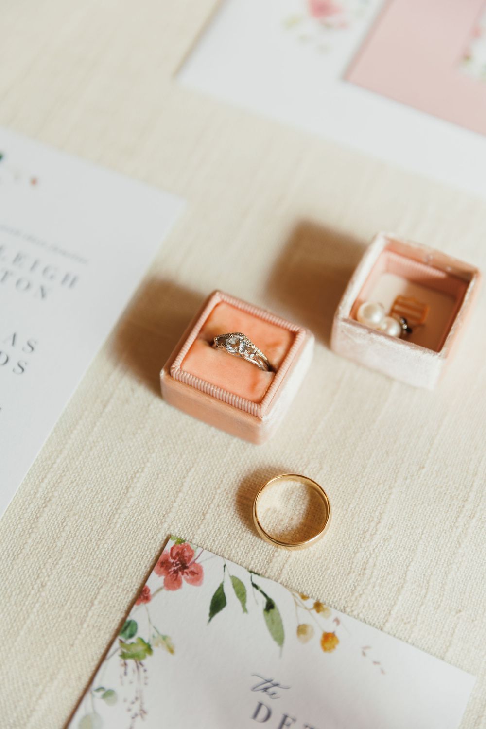 Delicate wedding rings displayed in peach-colored velvet ring boxes with floral stationery.