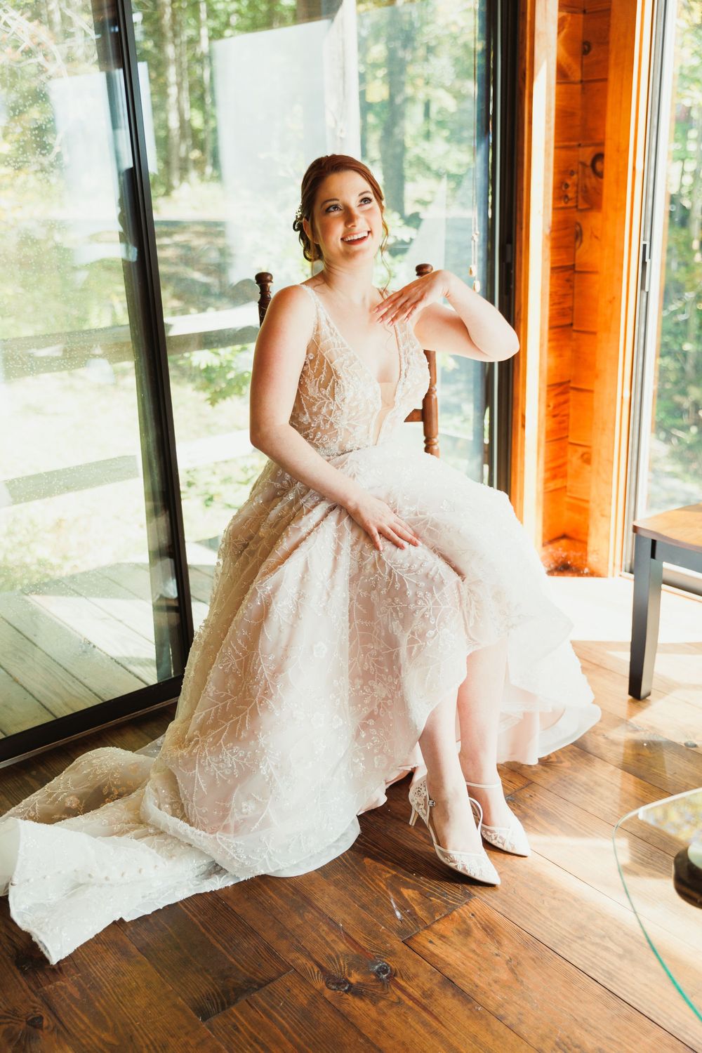A joyful moment captured of a bride in a lace gown sitting by a window in natural light.