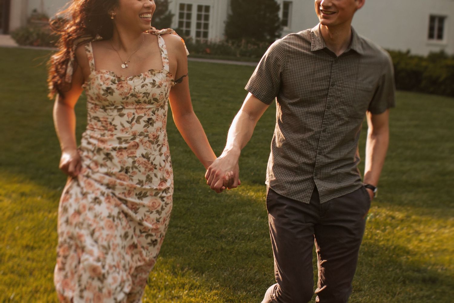 A couple walks hand in hand through a sunlit meadow, her floral dress flowing in the breeze.