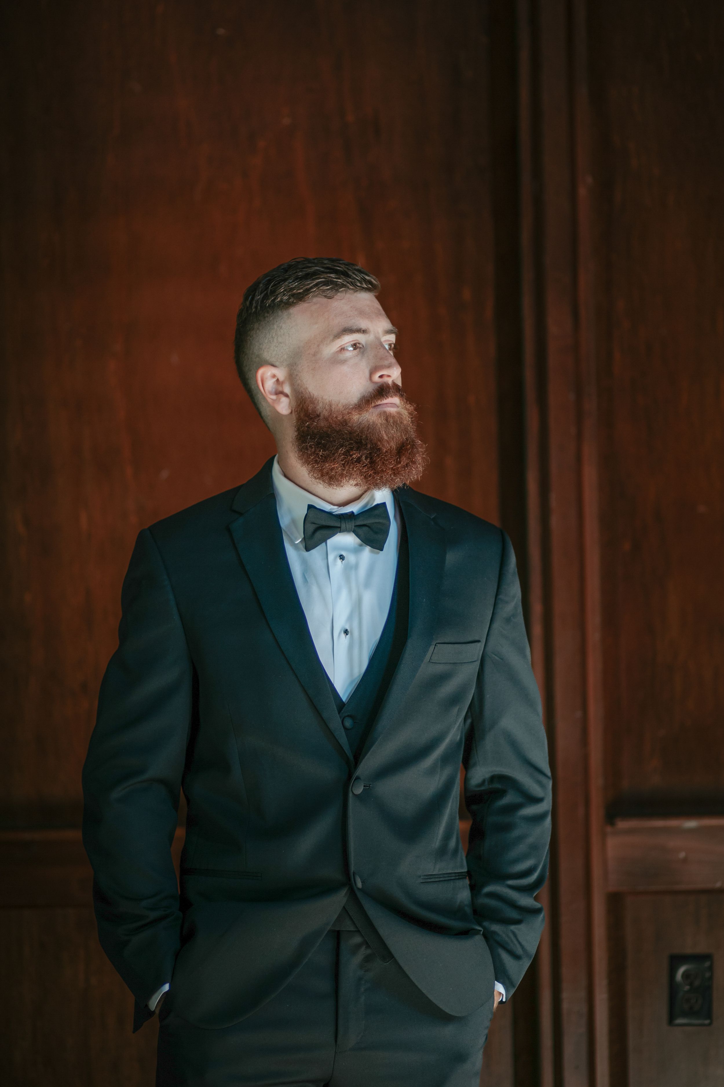 Groom in black tuxedo and bowtie poses for formal portrait against dark wood paneled wall.