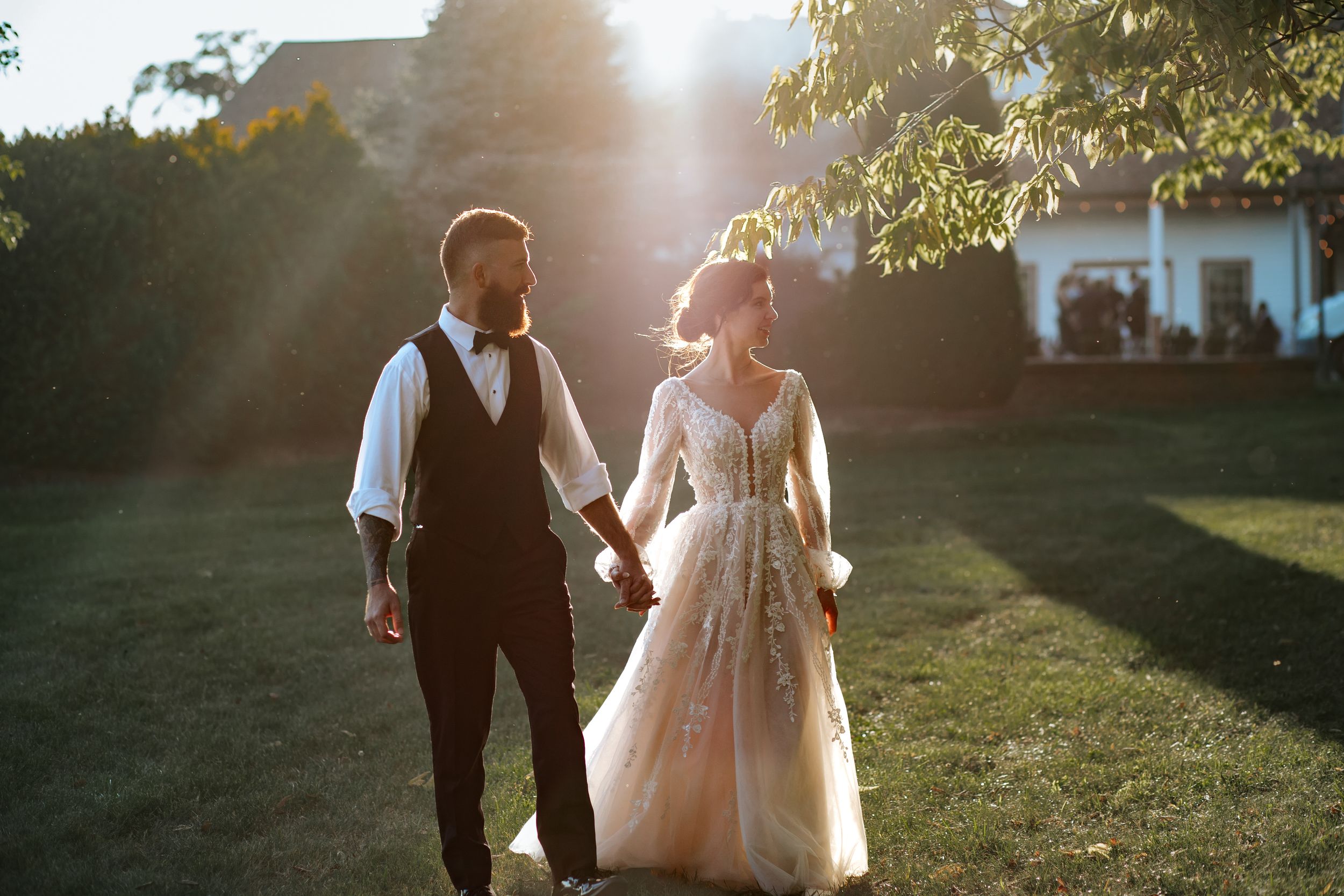 Wedding couple strolls through a garden backlit by warm evening sunlight.