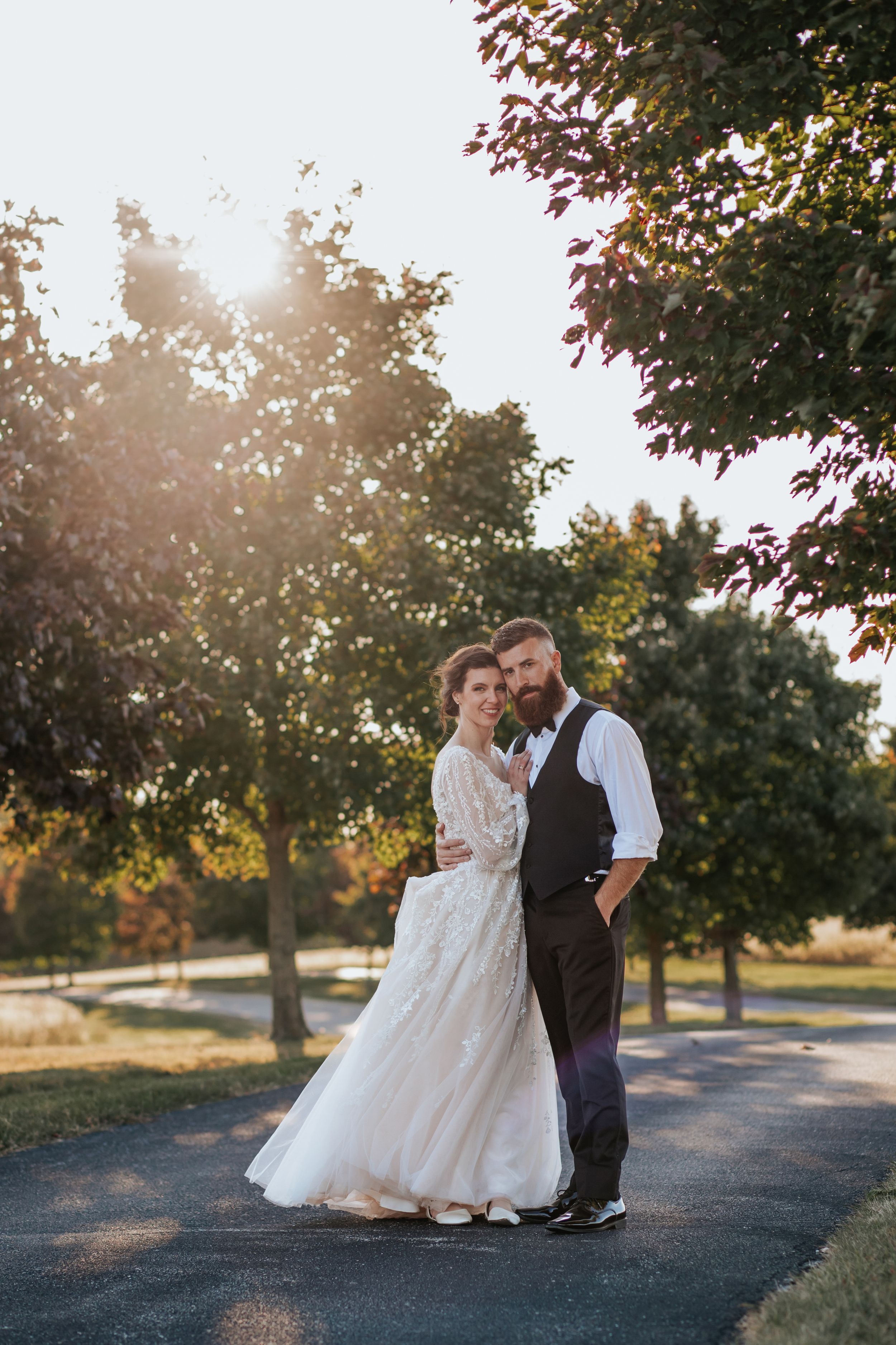 A romantic sunset wedding portrait shows a couple embracing on a tree-lined path with golden sunlight filtering through.