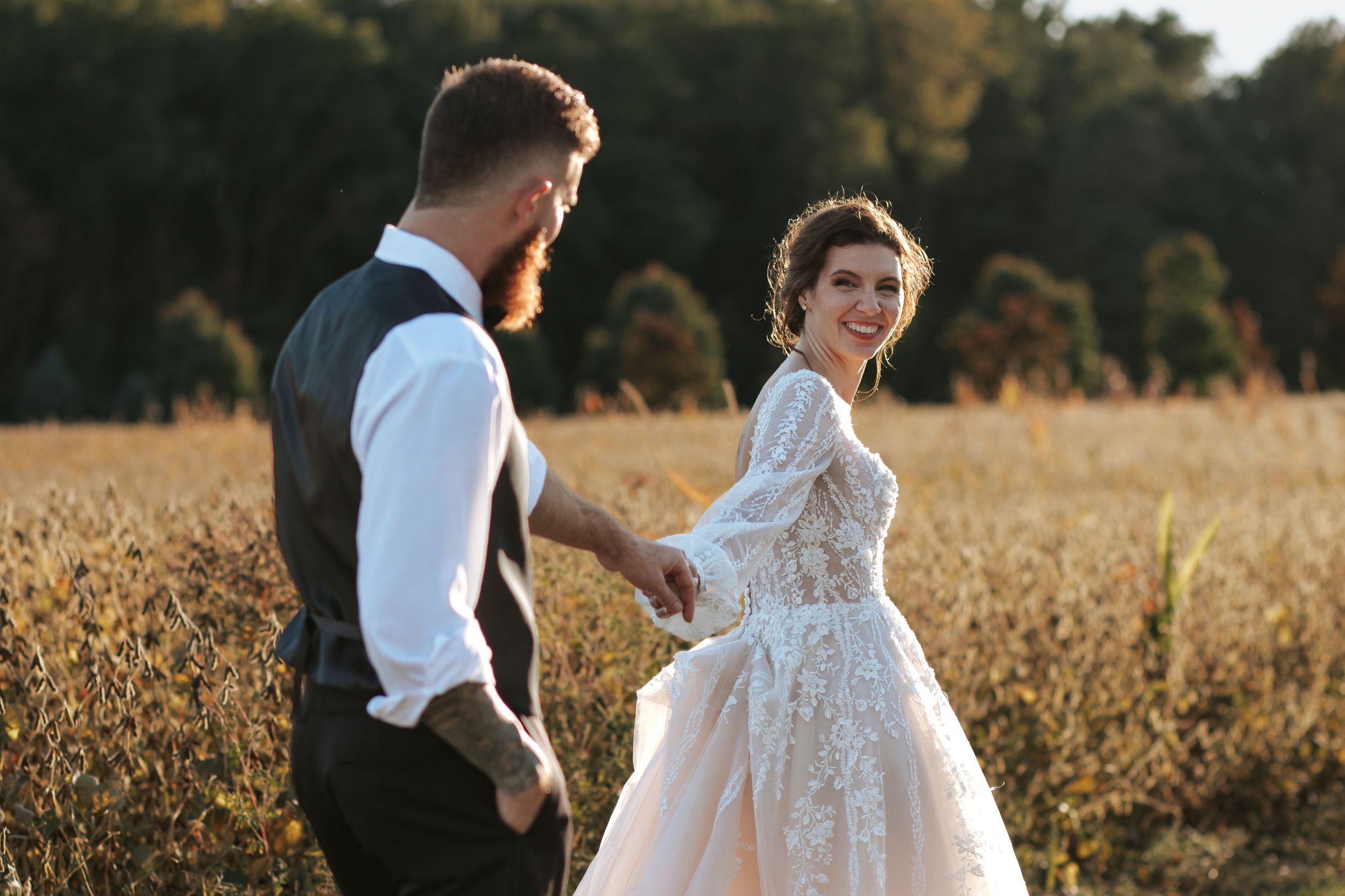 Wedding couple poses in a sunlit field wearing formal attire with bride in lace dress.