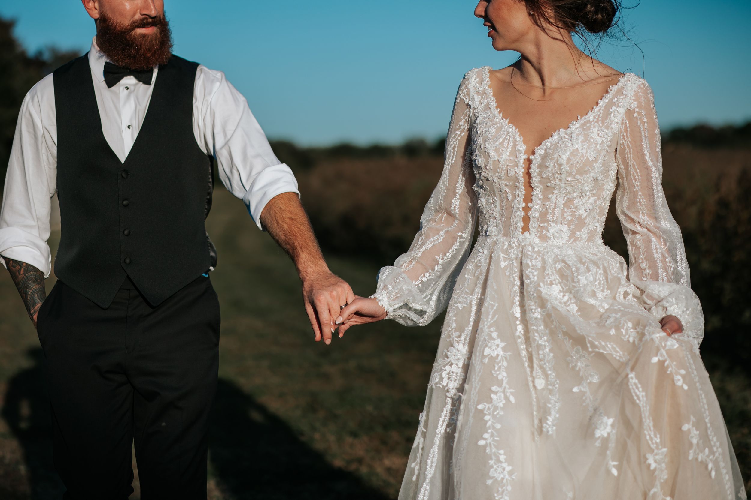 Wedding couple holds hands while walking through field, bride wearing flowing lace gown.