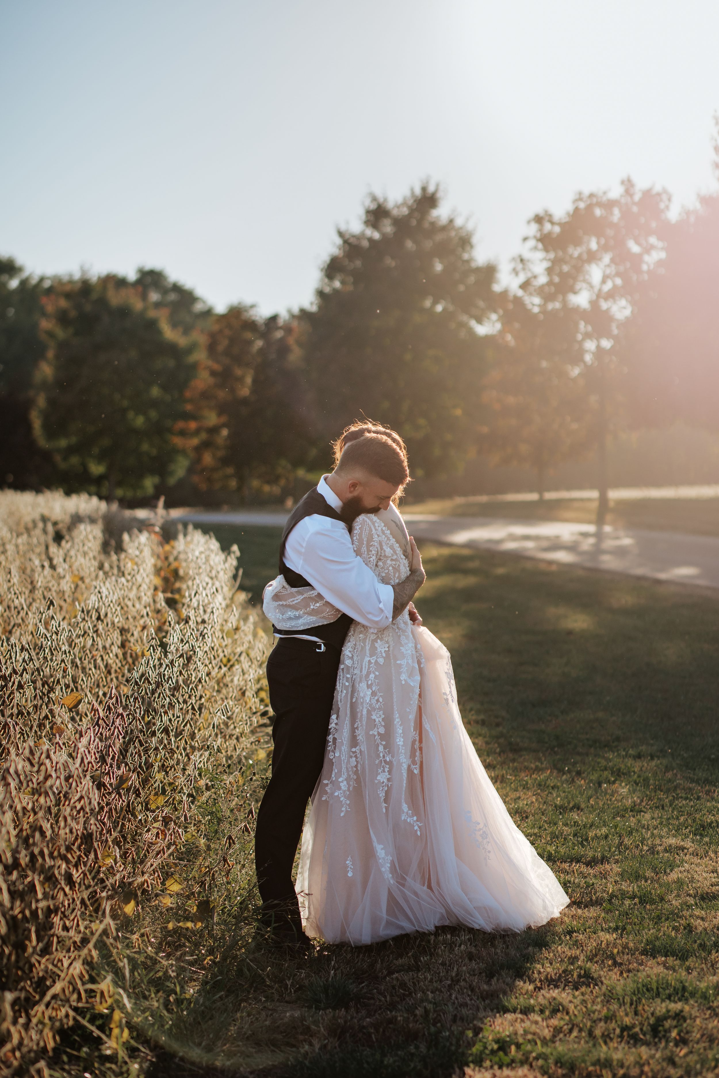 Backlit wedding portrait shows couple embracing in golden sunset light near ornamental grasses.