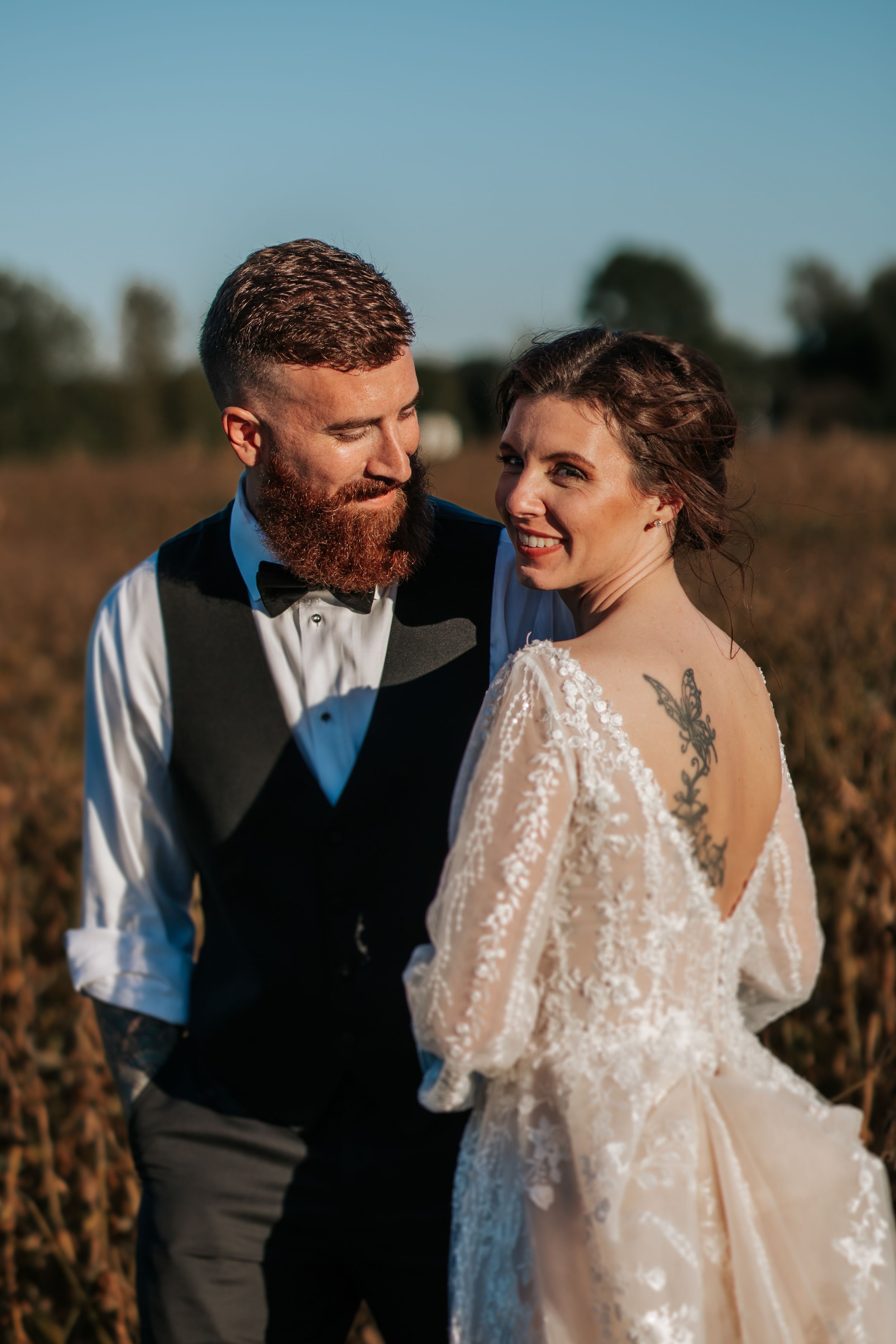 Candid wedding portrait in rustic field shows couple sharing joyful moment in formal attire.