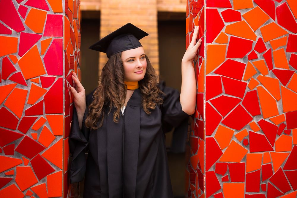 Graduates in black caps and gowns stand against a vibrant red and orange mosaic wall during commencement.