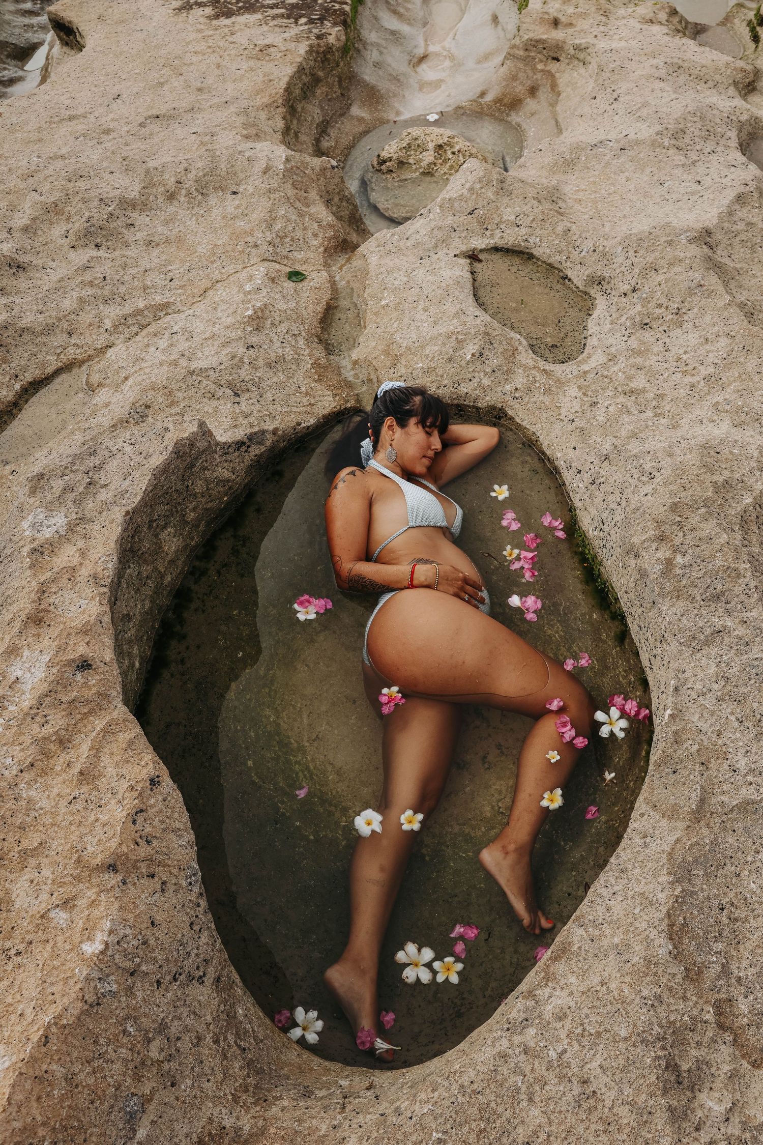 Woman in white bikini lying in a natural rock pool surrounded by tropical flowers on a beach.