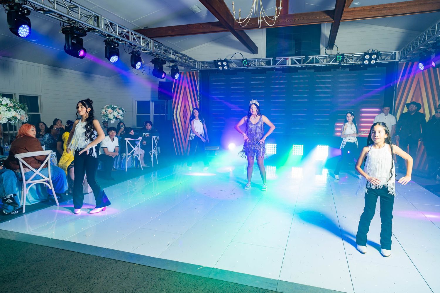 Performers on a white stage with blue lighting and theatrical spotlights during an indoor entertainment event.