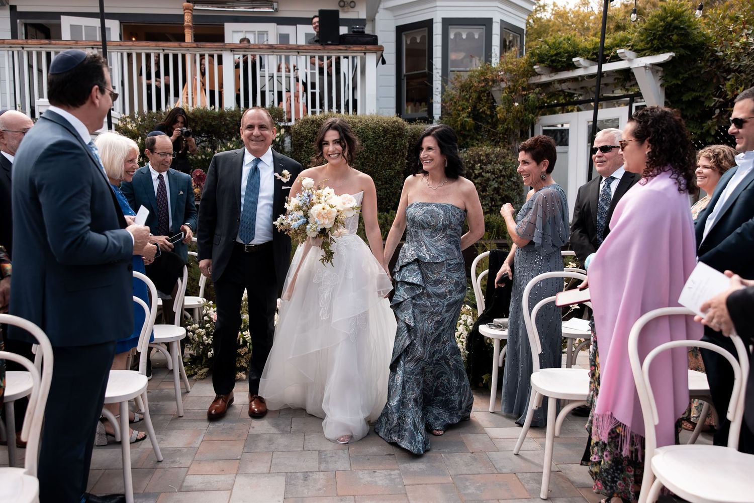 A wedding party walks through a rustic outdoor venue with white buildings and lush greenery in the background.