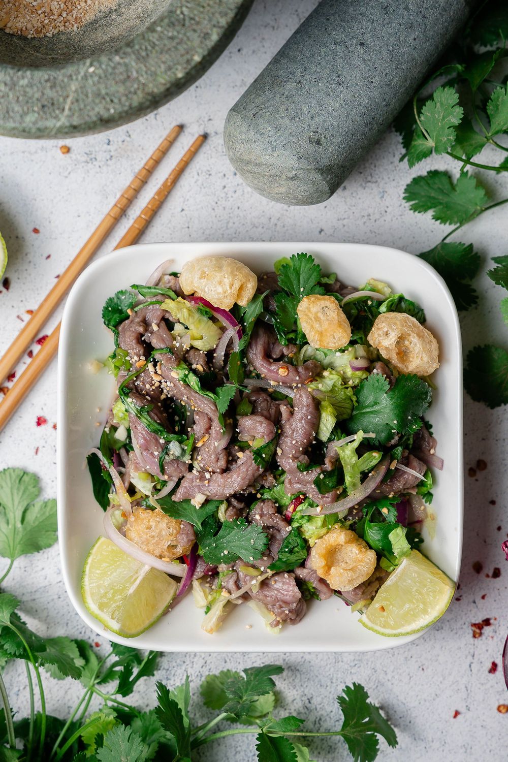 Grilled beef slices garnished with fresh cilantro and lime wedges served on a white rectangular plate with chopsticks.