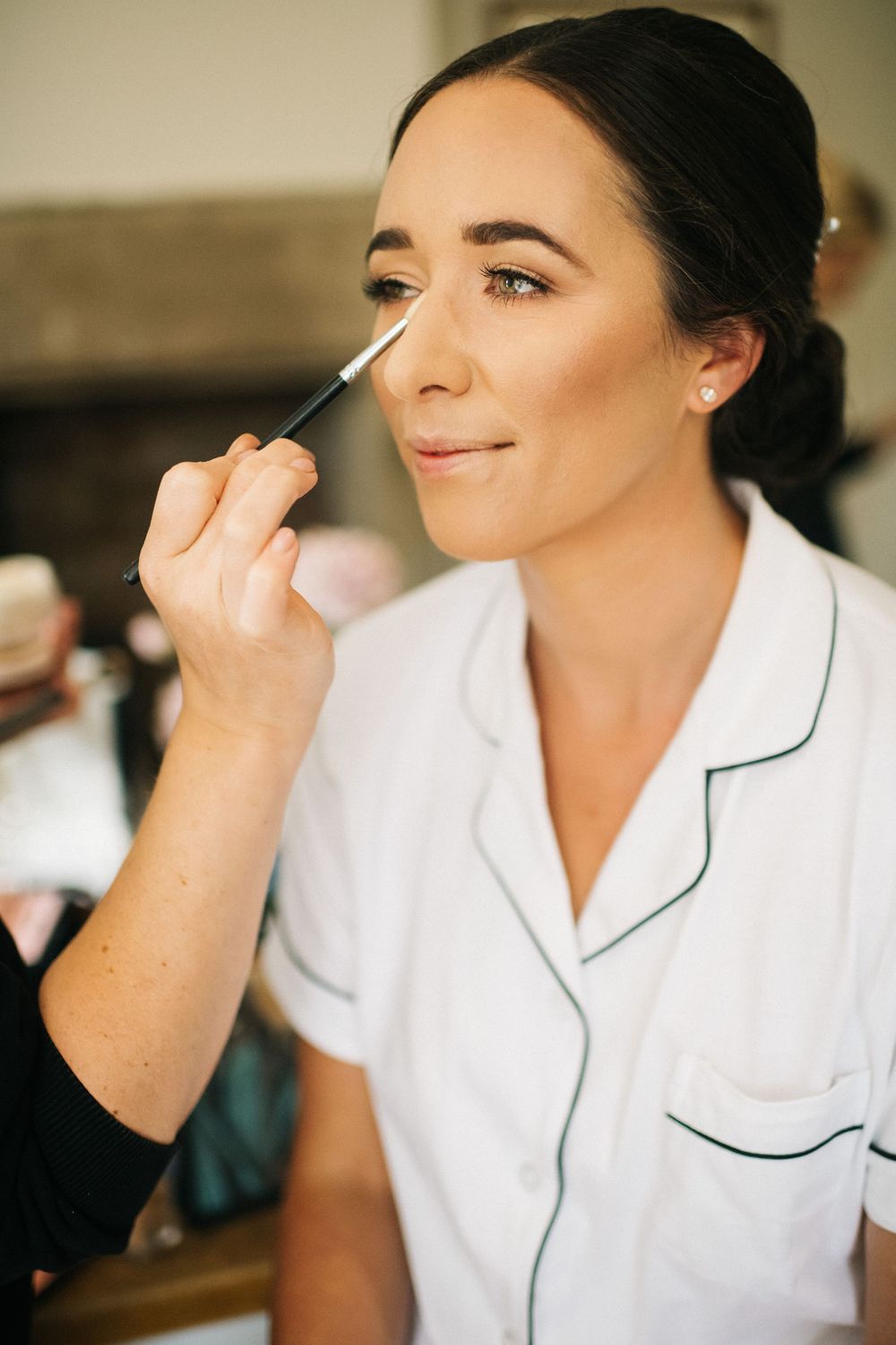 A makeup artist applies concealer with a brush while wearing a white uniform with navy piping.