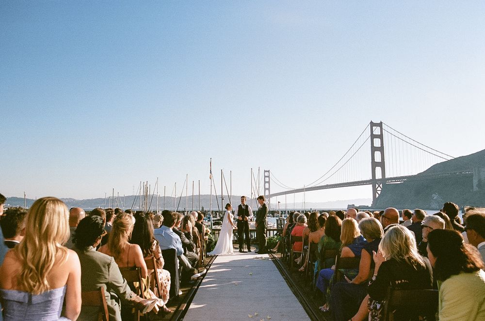 Outdoor wedding ceremony on a pier with the iconic Golden Gate Bridge visible in the background on a clear sunny day.