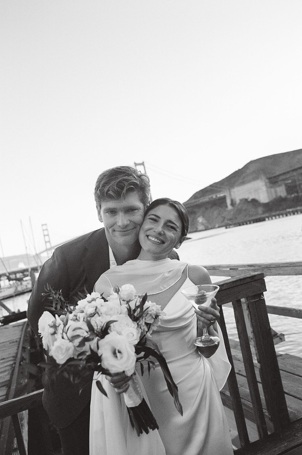 A black and white wedding photo showing a couple embracing on a boat with the Golden Gate Bridge in the background.