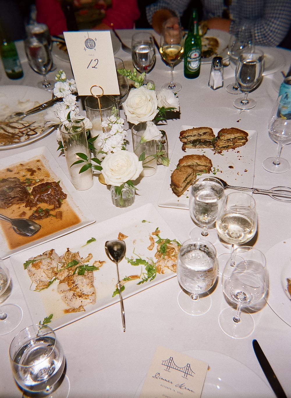 Wedding reception table settings with white flowers, wine glasses, and scattered food remnants on a white tablecloth.