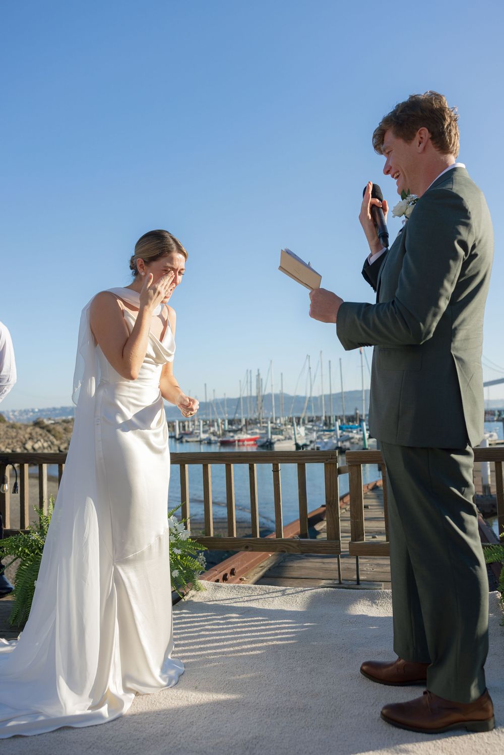A wedding ceremony takes place on a deck overlooking a marina with boats in the background on a sunny day.