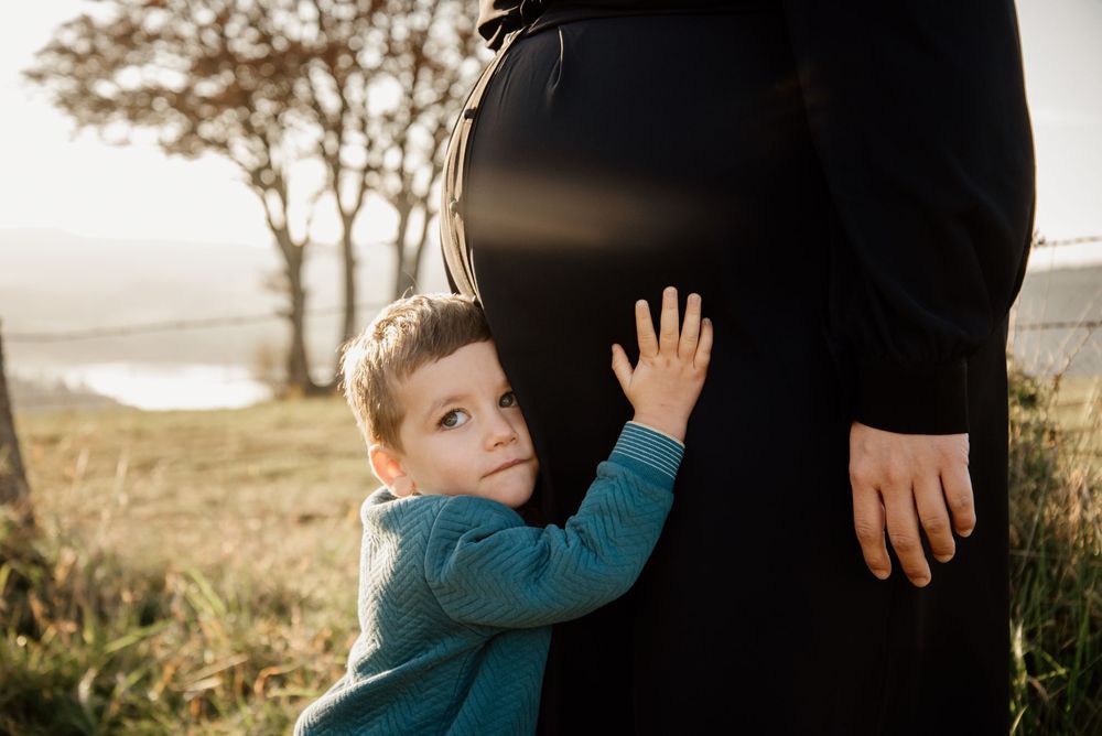 Young boy hugging a pregnant woman's belly outdoors in a scenic countryside setting.