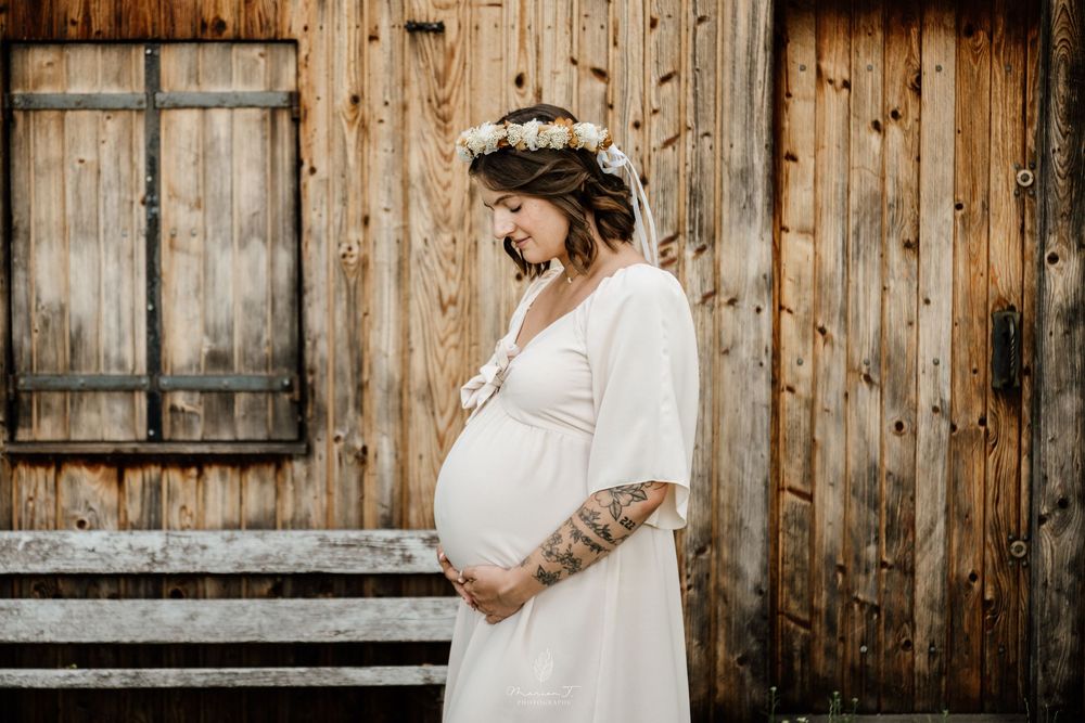 Tattooed pregnant woman in white dress with floral crown standing beside a rustic wooden wall.