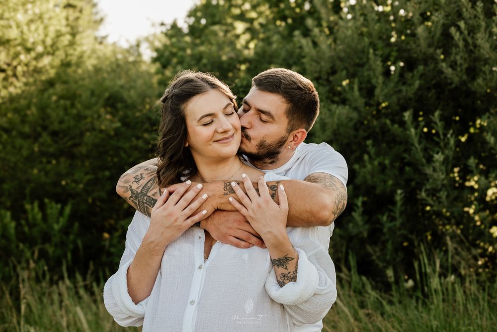 Tattooed couple embracing and kissing in a lush green outdoor setting in warm sunlight.
