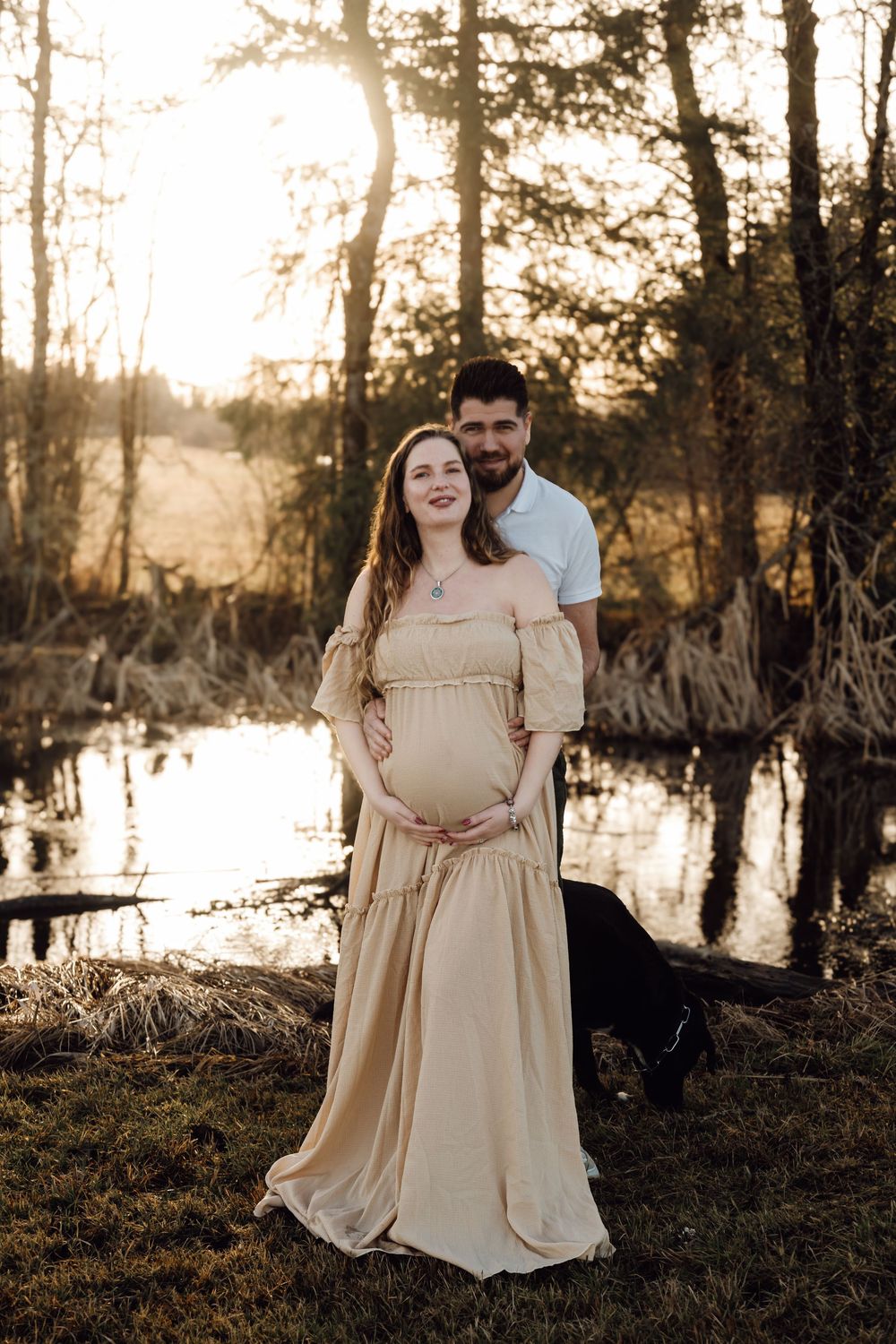 Couple embracing outdoors at sunset, pregnant woman in beige gown with partner holding her belly.