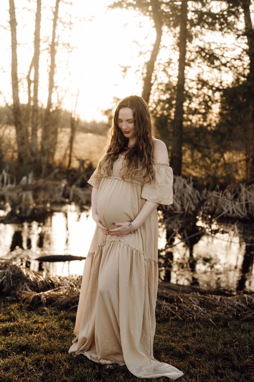 Pregnant woman in a flowing beige dress cradling her belly near a serene pond at sunset.