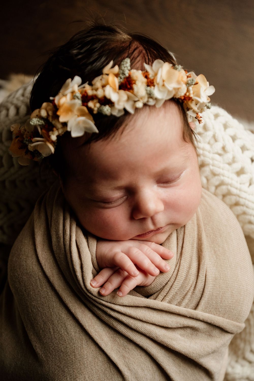 Sleeping newborn baby wrapped in beige swaddle wearing a delicate floral crown headband.