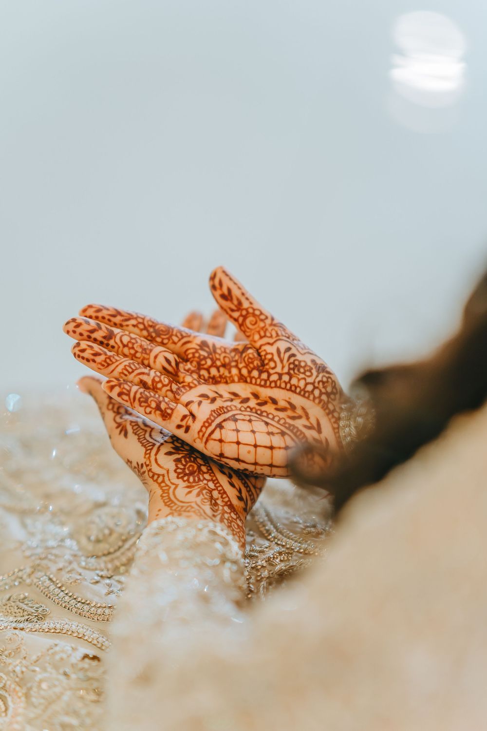 Intricate henna mehndi design on hands with traditional Indian bridal jewelry against white lace background.