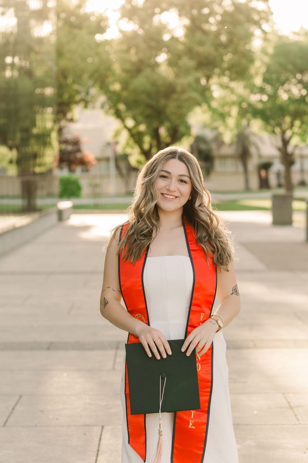 Graduate wearing orange and white graduation stole holds diploma while standing on campus walkway during sunset.