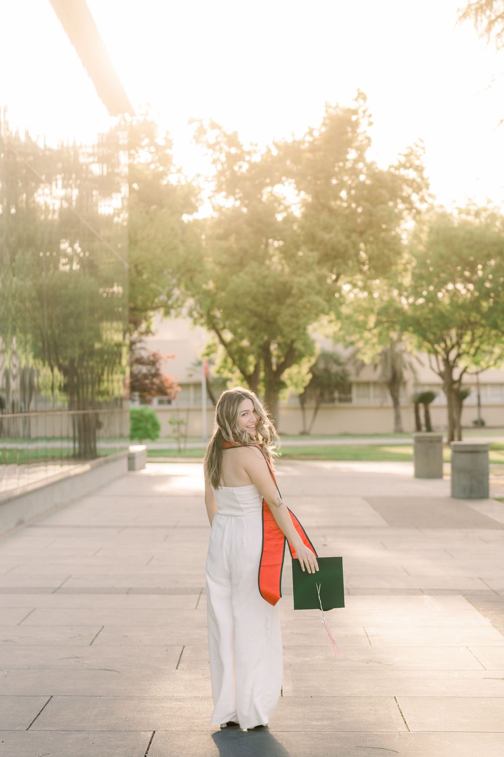 Graduate in white dress walking outdoors with coral bag during golden hour sunset on campus.