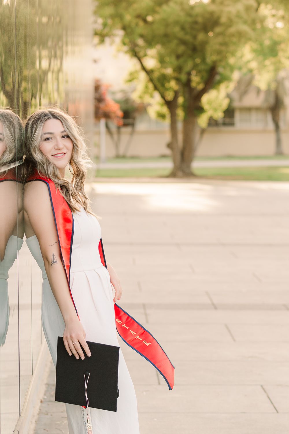 Graduate posing with diploma and sash against a sunny outdoor campus backdrop with trees.