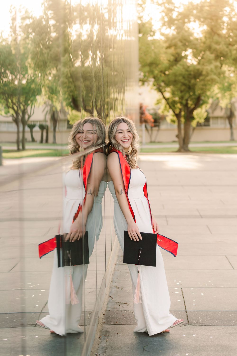 Graduate in white dress and red stole poses with reflection in glass window on sunny graduation day.